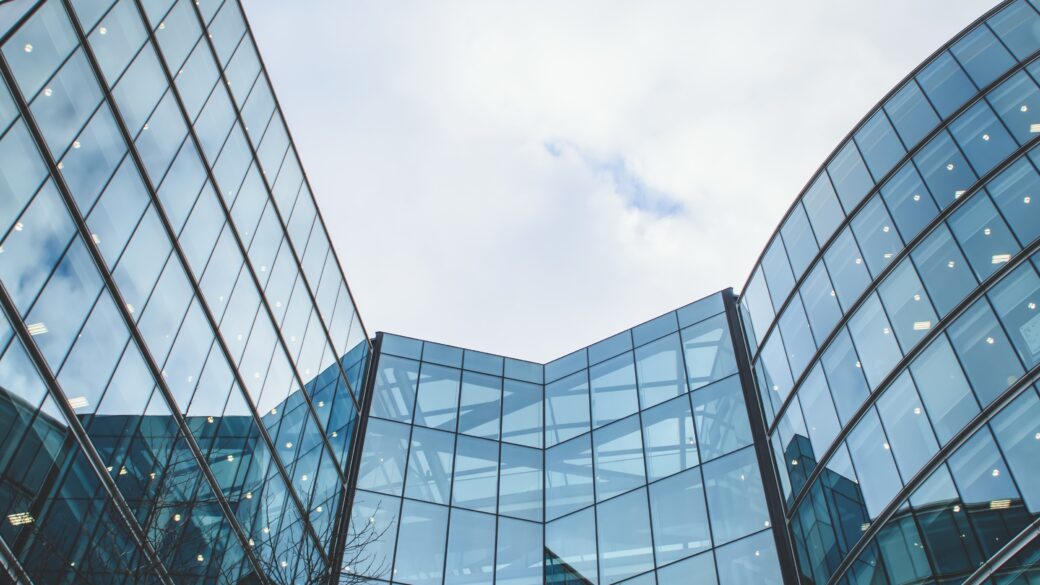A modern office building stands strongly, its glass exterior reflecting a blue sky and white clouds.